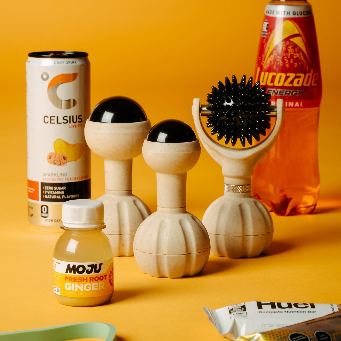 A set of handheld massager and with black spherical tops and a black spiky ball, arranged on a bright yellow-orange studio background alongside branded beverage packaging including Celsius, Lucozade, and Moju.