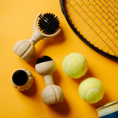 A styled flat-lay of massage tools and accessories on a bright orange background, including a tennis ball, and part of a tennis racket.