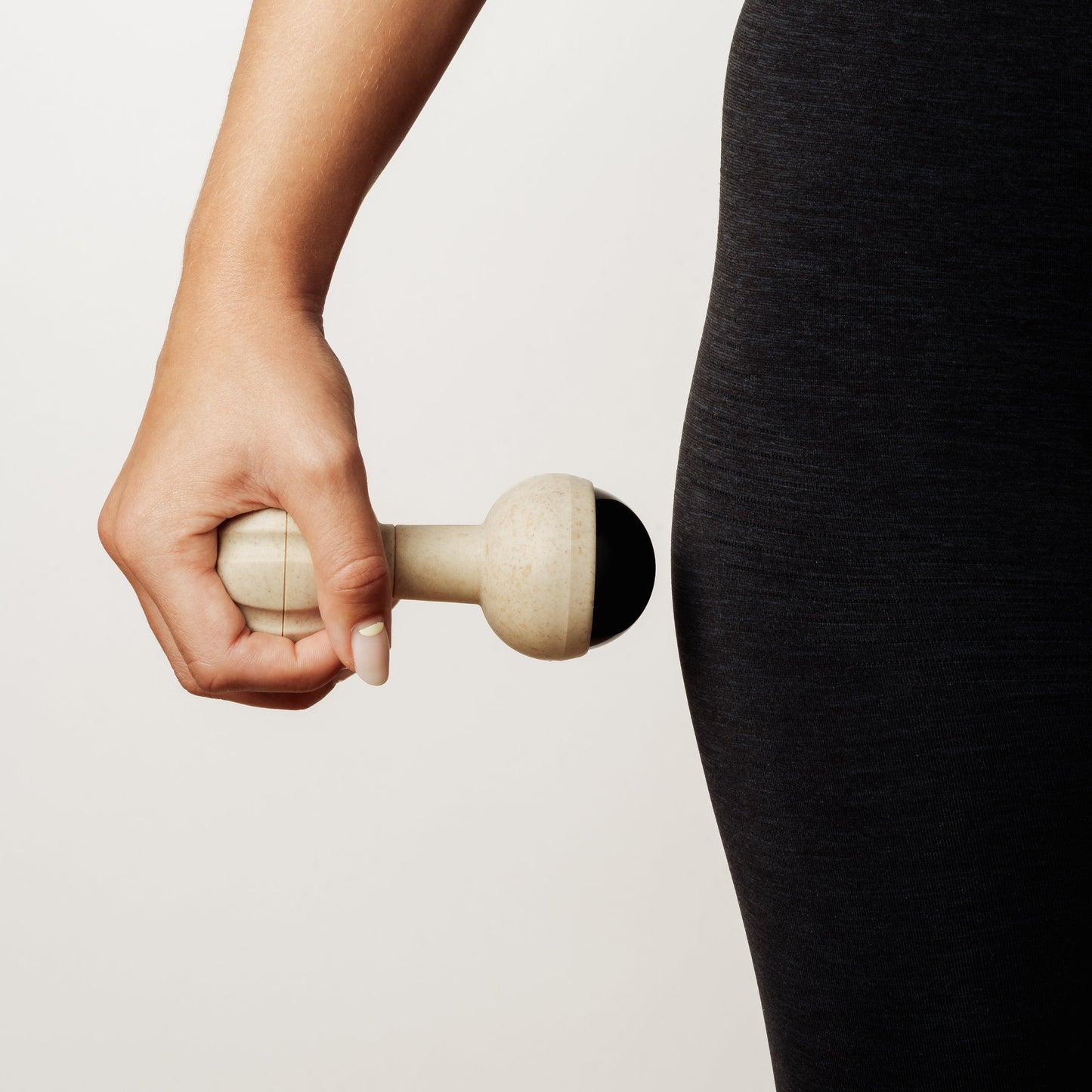 A close-up of a person's hand holding a rollerball massager against their hip in black leggings, set against a clean off-white background.