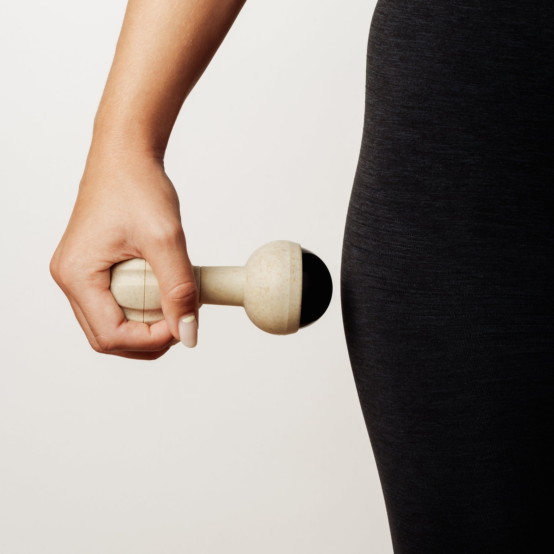 A close-up of a person's hand holding a rollerball massager against their hip in black leggings, set against a clean off-white background.