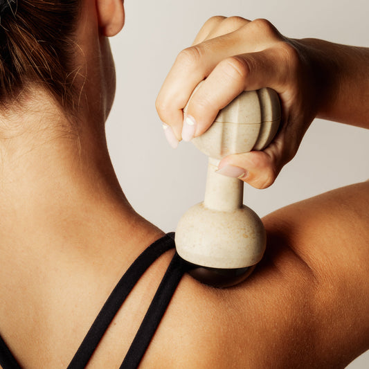 A close-up lifestyle shot of a person using a beige handheld, rollerball massage tool on their shoulder, showing the hand pressing the tool onto the skin.