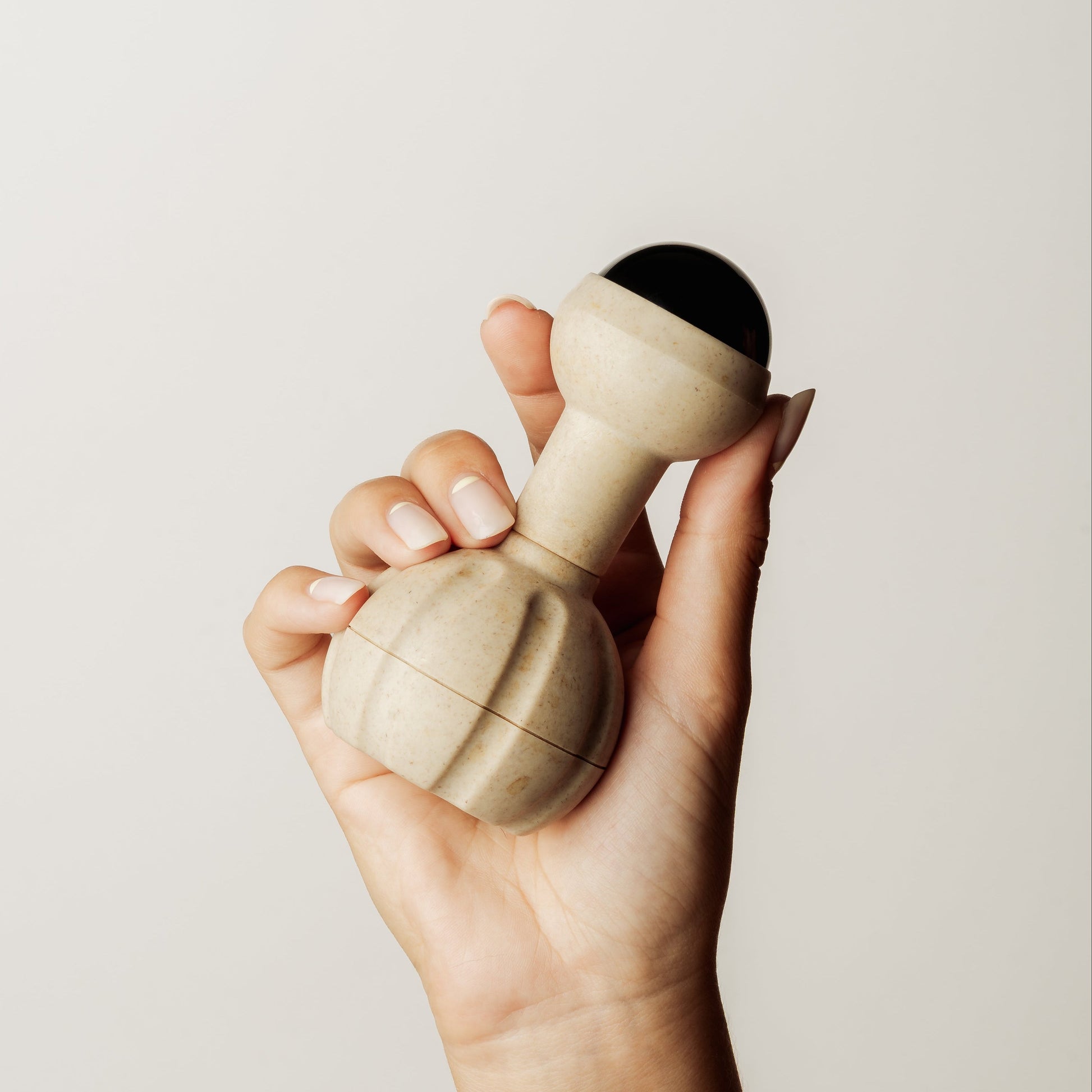 A hand holding a natural coloured handheld massager with a black rollerball head, set against a plain light gray background.