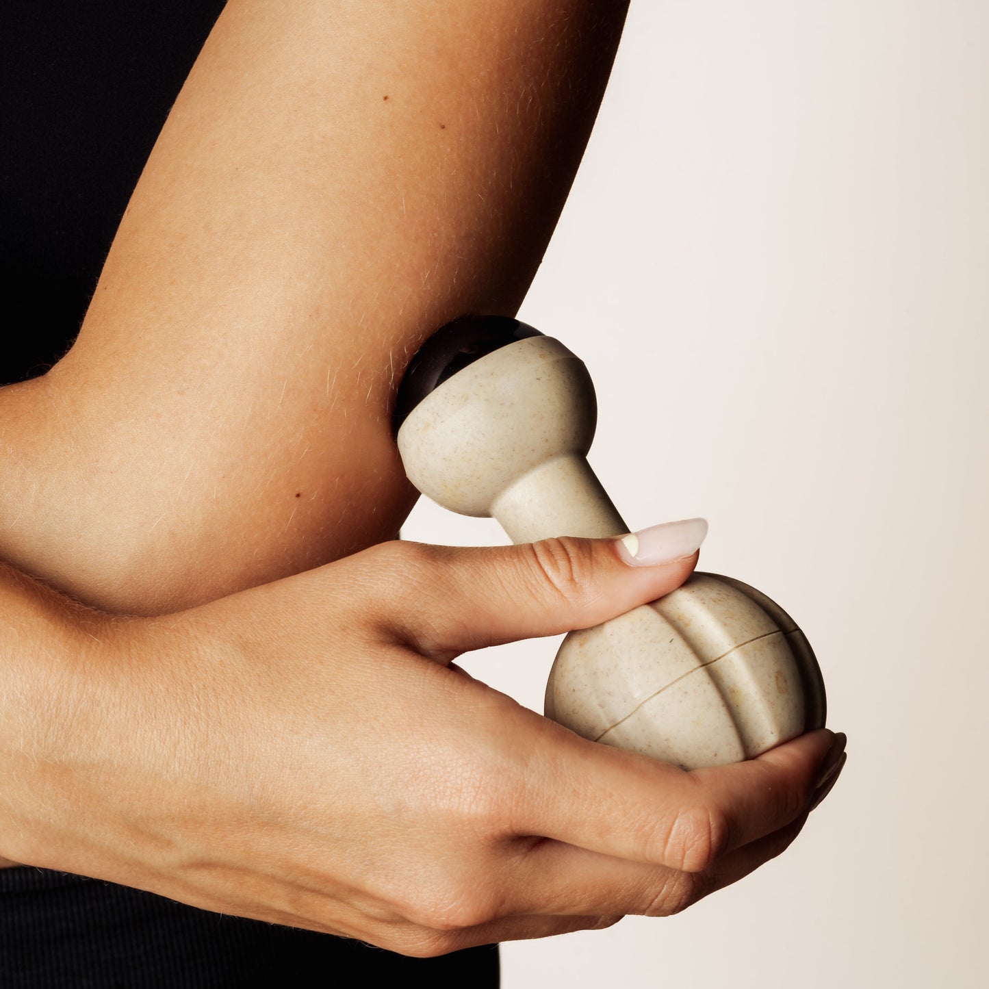 A close-up of a person using a handheld rollerball massage tool on their upper arm. The tool has a rounded, beige handle with a black rollerball, and the person's hand and forearm are visible against a plain light background.