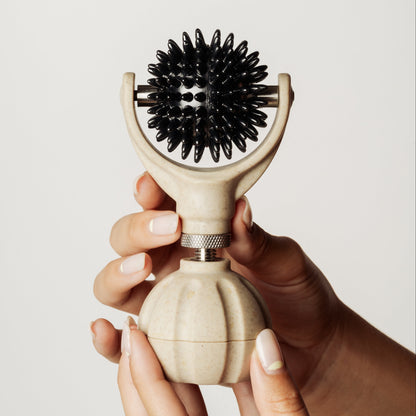 A close-up of a person's hands holding a handheld massager. The massager features a round beige handle and a black spikey ball with a central knurled metal adjustment collar.