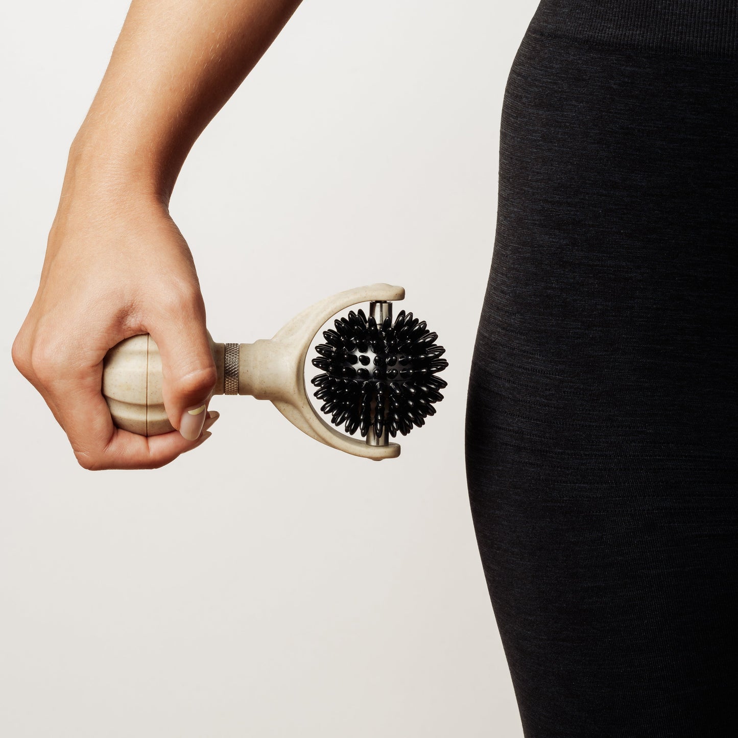 A close-up of a person holding a handheld black-and-beige handheld spikey roller against their hip, wearing dark gray leggings, on a clean off-white background.