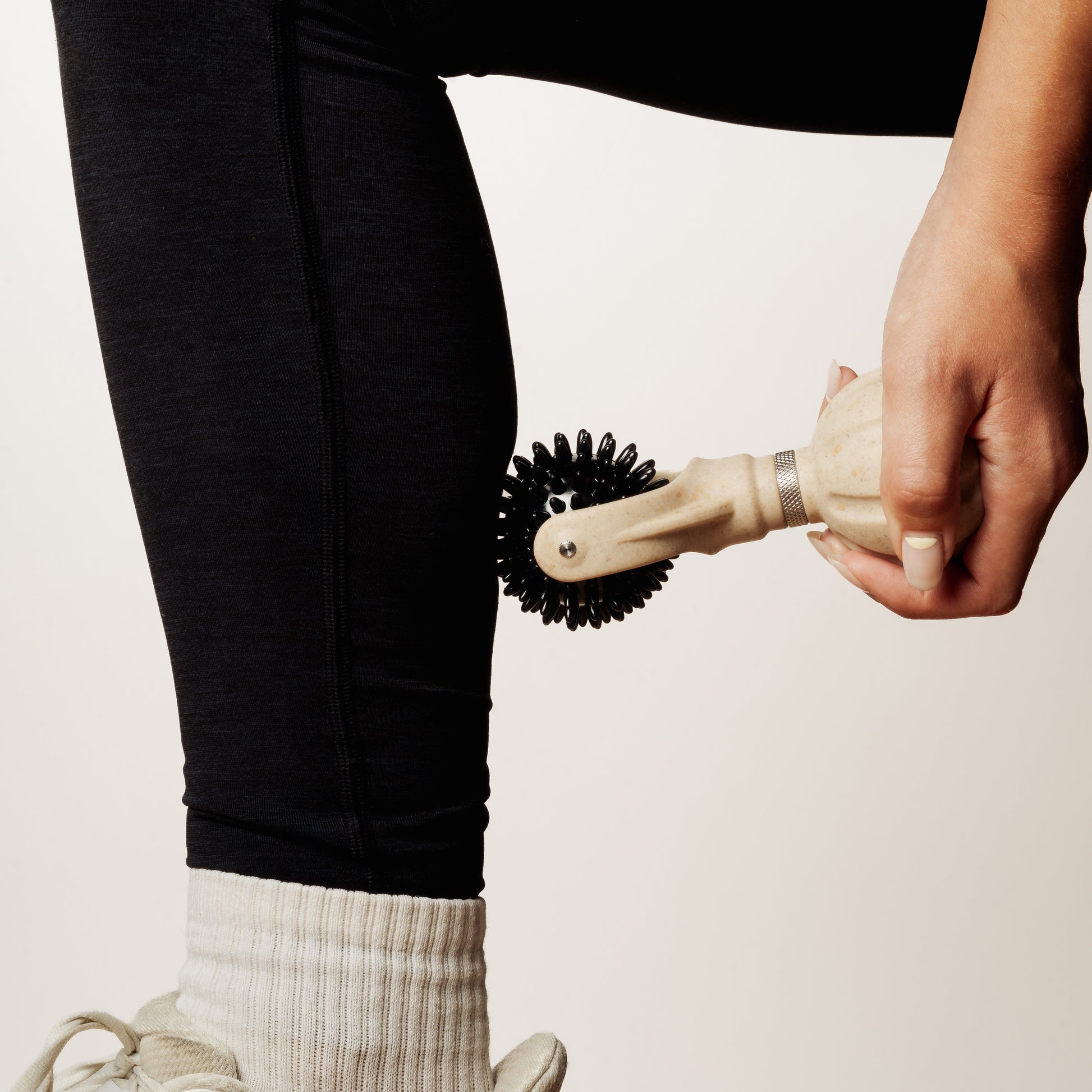 A close-up of a person's leg wearing black leggings and a white sock, holding a handheld black-and-beige spikey ball roller against the calf.