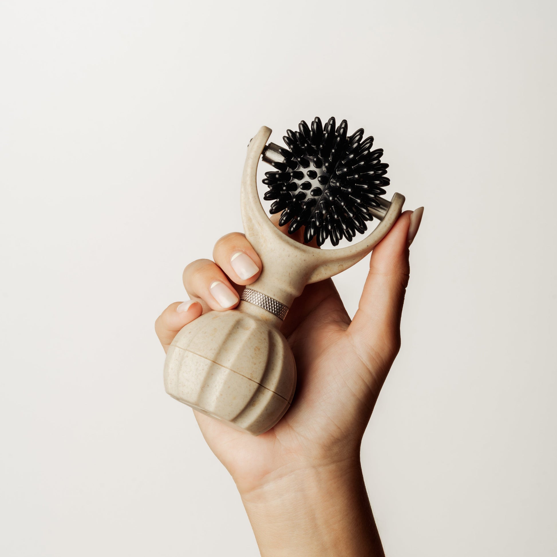 A hand holding a handheld massager with a beige handle and black silicone bristles against a plain off-white background.