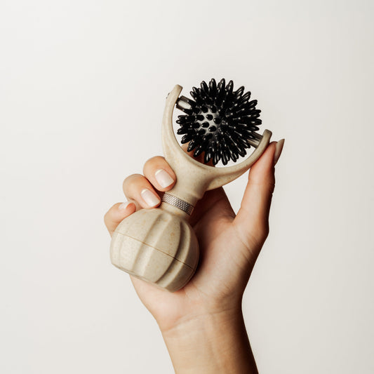 A hand holding a handheld massager with a beige handle and black silicone bristles against a plain off-white background.