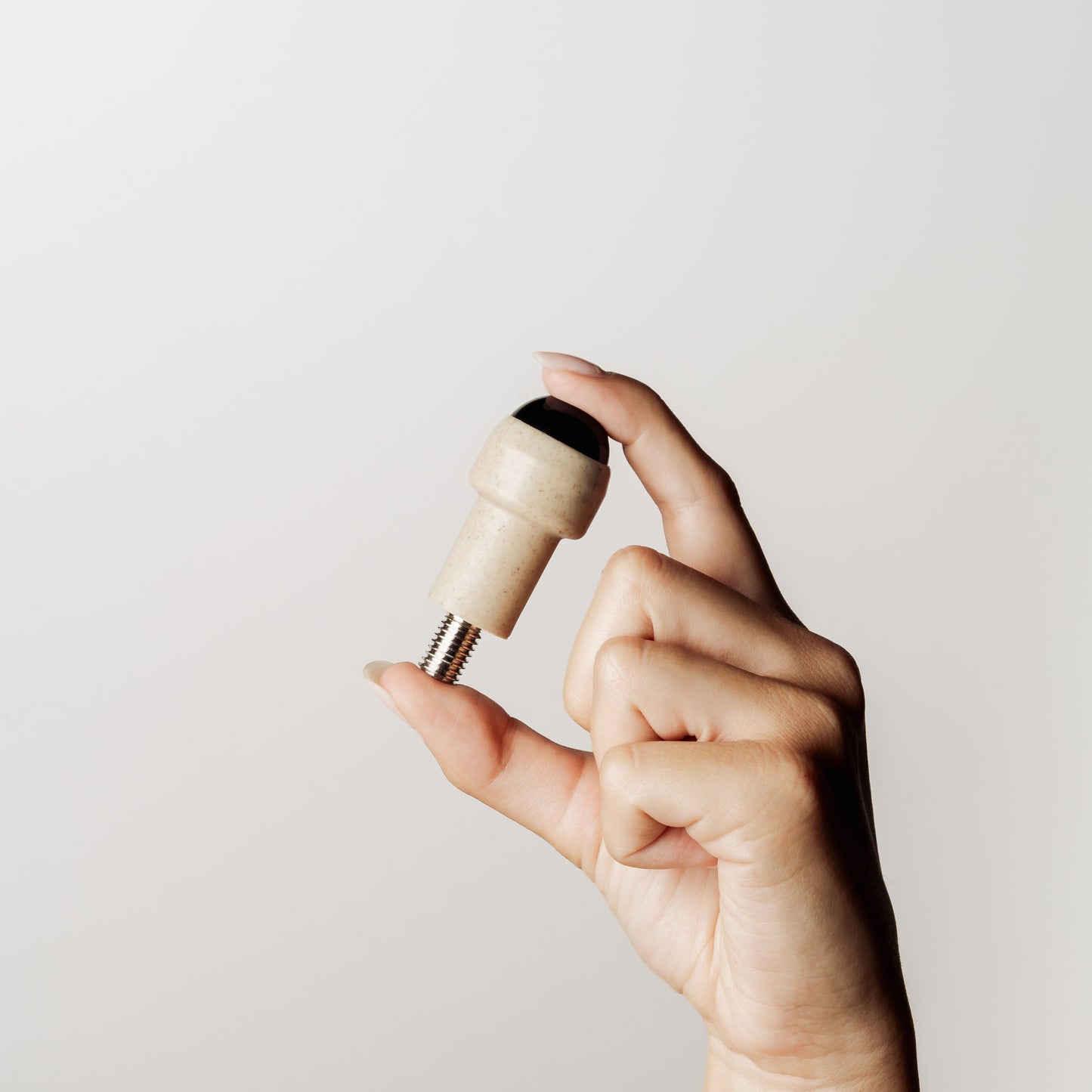 A hand holding a small beige cylindrical accessory with a black rollerball top and a threaded metal base against a plain light gray background.